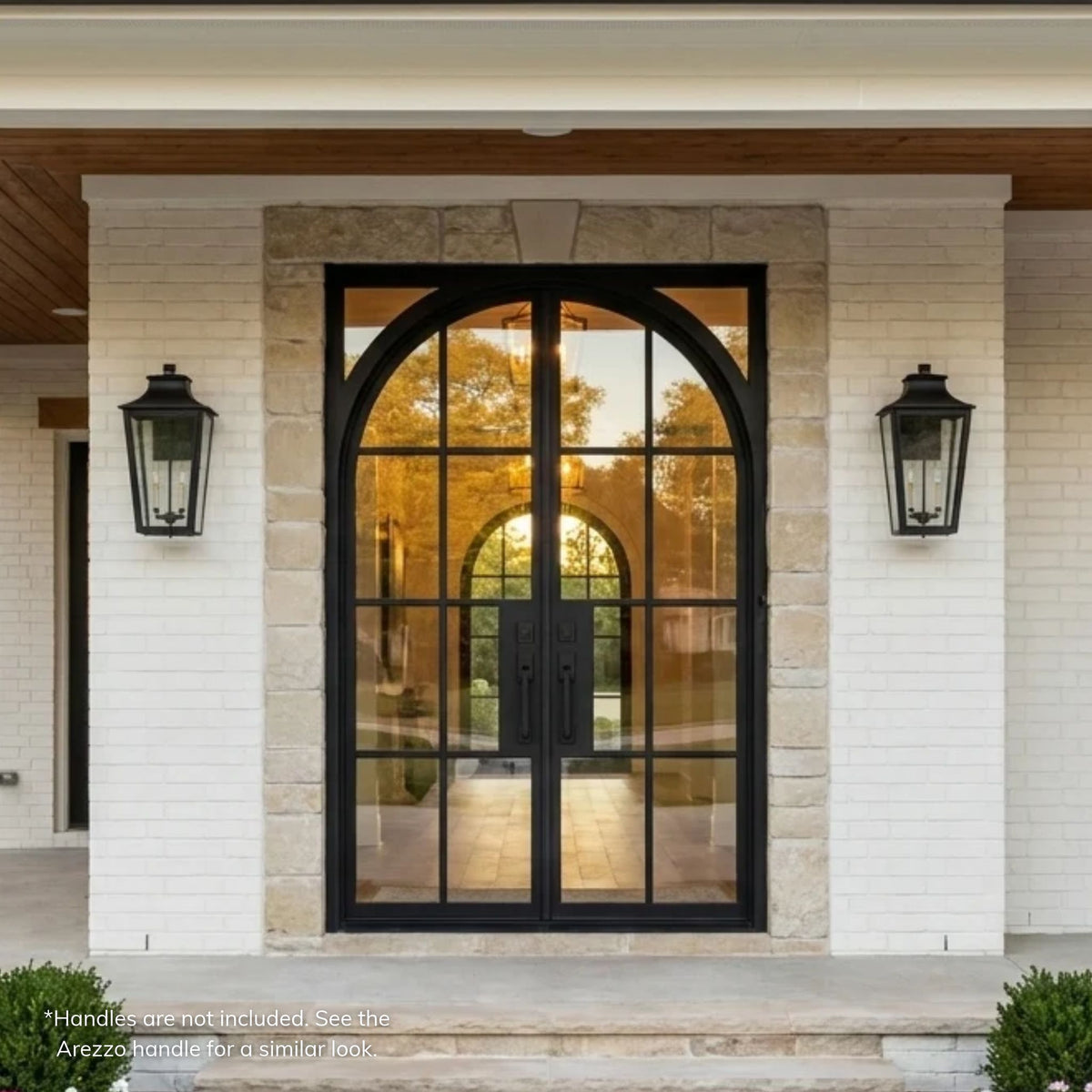 Geoffrey black wrought iron double entry door with arched clear glass grid design, set in a cut stone surround on a white brick home with a covered front porch