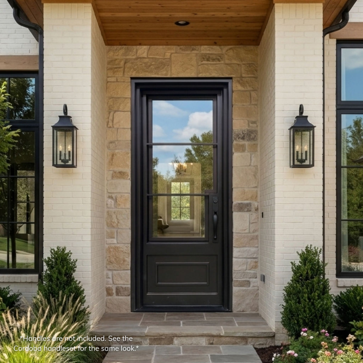 Wrought iron single entry door in matte black finish with a 3-lite clear glass upper section over a solid raised panel lower section, installed on a stone and brick traditional-style home with lantern sconces.